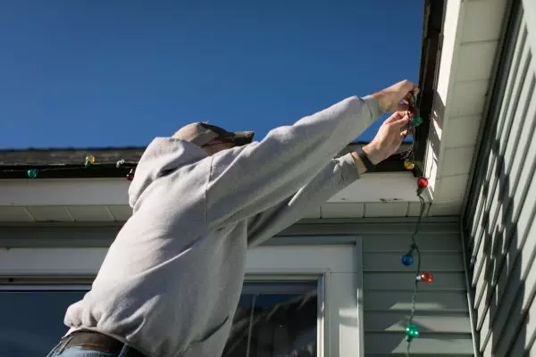 man hanging holiday lights on gutter of house