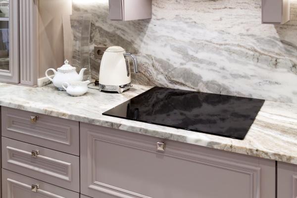 beautiful closeup of custom designed kitchen, with marble looking quartz countertop and backsplash. cream electric kettle with porcelain tea accessories on the marble countertop next to ceramic hob