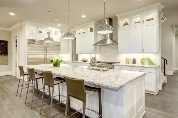 White kitchen design features large bar style kitchen island with granite countertop illuminated by modern pendant lights. Stainless steel appliances framed by white shaker cabinets and cabinets above the regular cabinets.